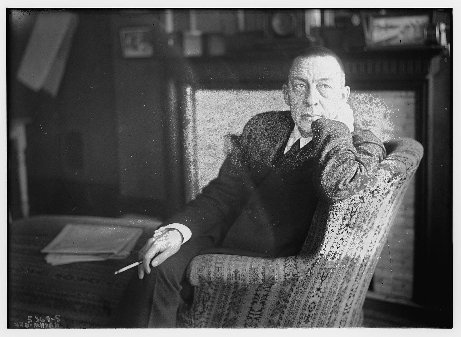 A black and white photo of the composer Rachmaninoff sitting in a chair, resting his head against his hand, with a cigarette in the other, his expression pensive.