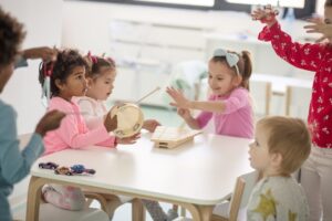 Preschool children playing musical instruments such as a tamborine, an xylophone, the triangle, and hand bells. One of the children, with curly black hair, looks up in wonder.