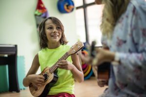 Boy playing the ukulele in school. He is looking up with a smile at his teacher, who is in the foreground, slightly blurring, but also playing the ukulele.