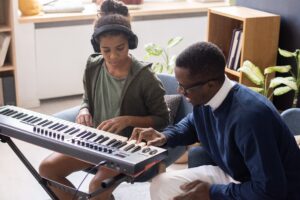 Boy sitting at keyboard, attentively listening to his male teacher, who demonstrates how to play the music.